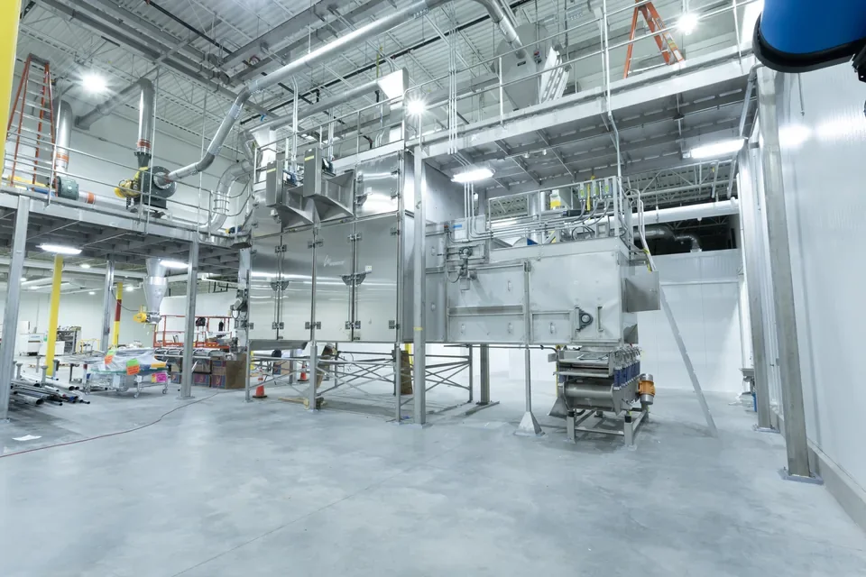 Stainless-steel processing equipment beneath overhead conduit and lighting in a production facility.