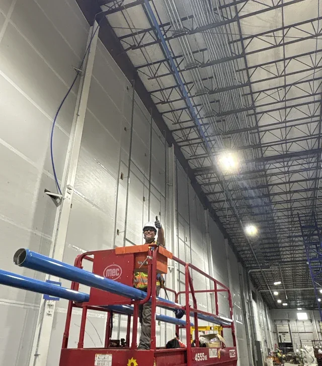 A plumbing installer on a red elevated work platform beside blue piping inside a distribution facility.
