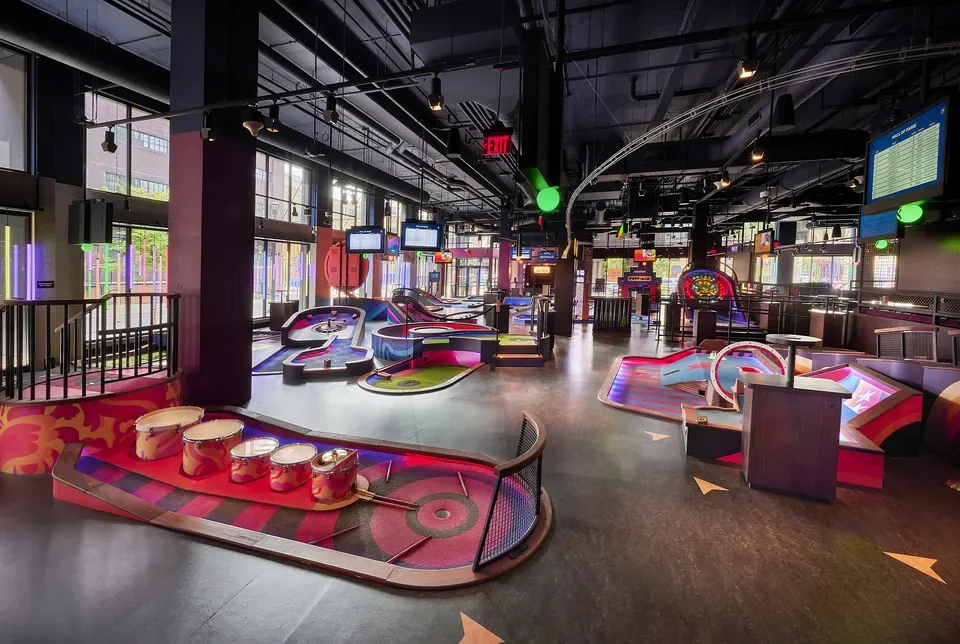 Colorful illuminated miniature golf features beneath exposed ceiling services.